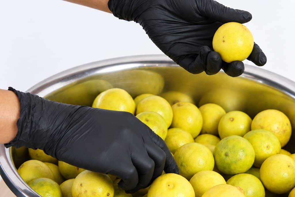 Gloved hands over a steel bowl of whole Cambodian limes, lifting one out to look at it.