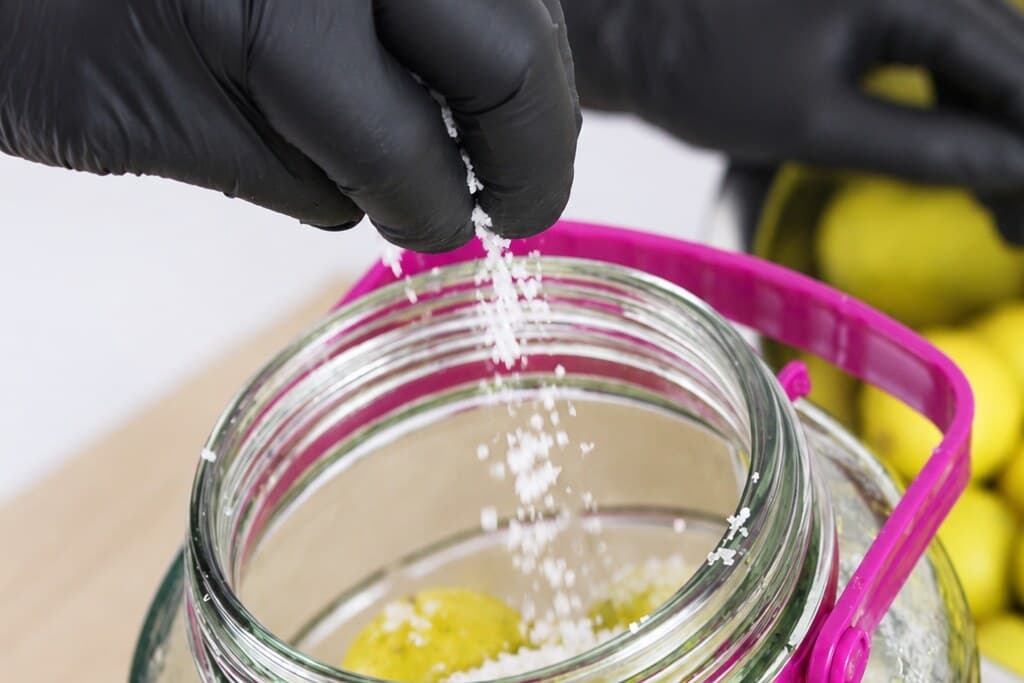 Sea salt falling from a gloved hand into the mouth of a tall jar, onto the limes packed below.