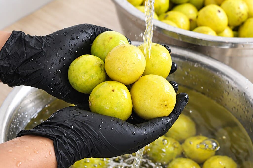 Water poured over a handful of limes held in gloved hands, above a bowl of fruit already in the water.