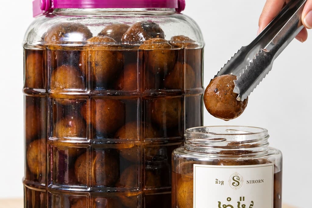 A cured lime held out of the jar in tongs, dark brown and whole, with the curing jar beside it.