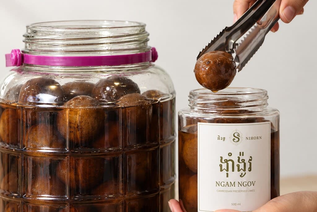 A cured lime lowered by tongs into a labelled 500 ml jar of Ngam Ngov held in one hand, the curing jar beside it.